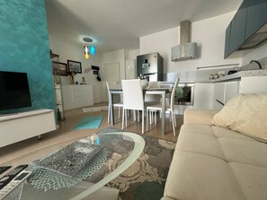 Un salon moderne avec un canapé, une table basse en verre et un téléviseur. On y trouve également un coin repas avec une table et des chaises. La cuisine est visible et comprend un réfrigérateur et une cuisinière.