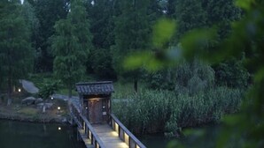 A wooden bridge leading to a traditional-style building with a tiled roof, surrounded by dense greenery and a calm body of water. - Yayunju Inn (Shanghai)