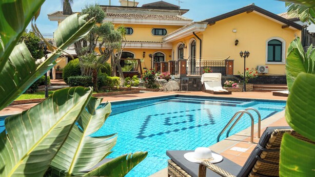 A swimming pool with a tiled deck, surrounded by lush greenery and a yellow building with a tiled roof. - Villa Gialla Private Padel Court (Aci Sant'Antonio)