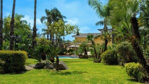 A well-maintained garden with a swimming pool, palm trees, and a building with a tiled roof. - Villa Gialla Private Padel Court (Aci Sant'Antonio)