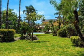 Un jardin bien entretenu, doté d’une piscine, de palmiers et d’un bâtiment dont le toit est recouvert de tuiles.