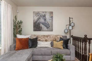A grey sofa with multiple cushions, a wall-mounted horse painting, a floor lamp, and a staircase with a wooden railing.