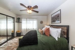 A bedroom with a bed, a ceiling fan, a window with curtains, a clock, and a framed picture on the wall.