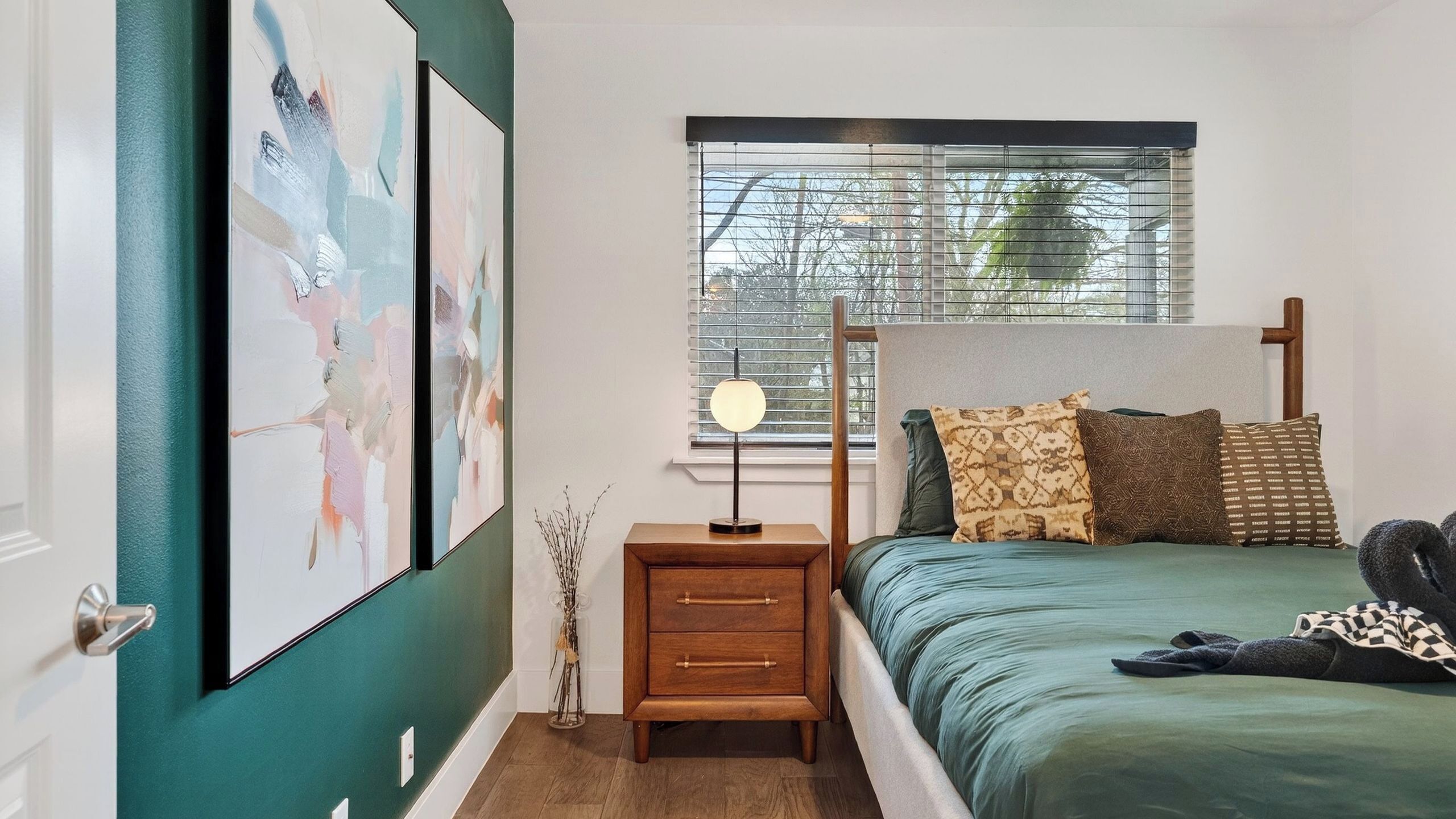 A bedroom with a bed, a bedside table with a lamp, a window with blinds, and artwork on the wall.