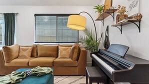 A living room with a sofa, a piano, a lamp, and a window with blinds. - Eastside Keys by Avantstay Bright East Austin Stay + Backyard Dining + Near 6th St and Downtown (Austin)