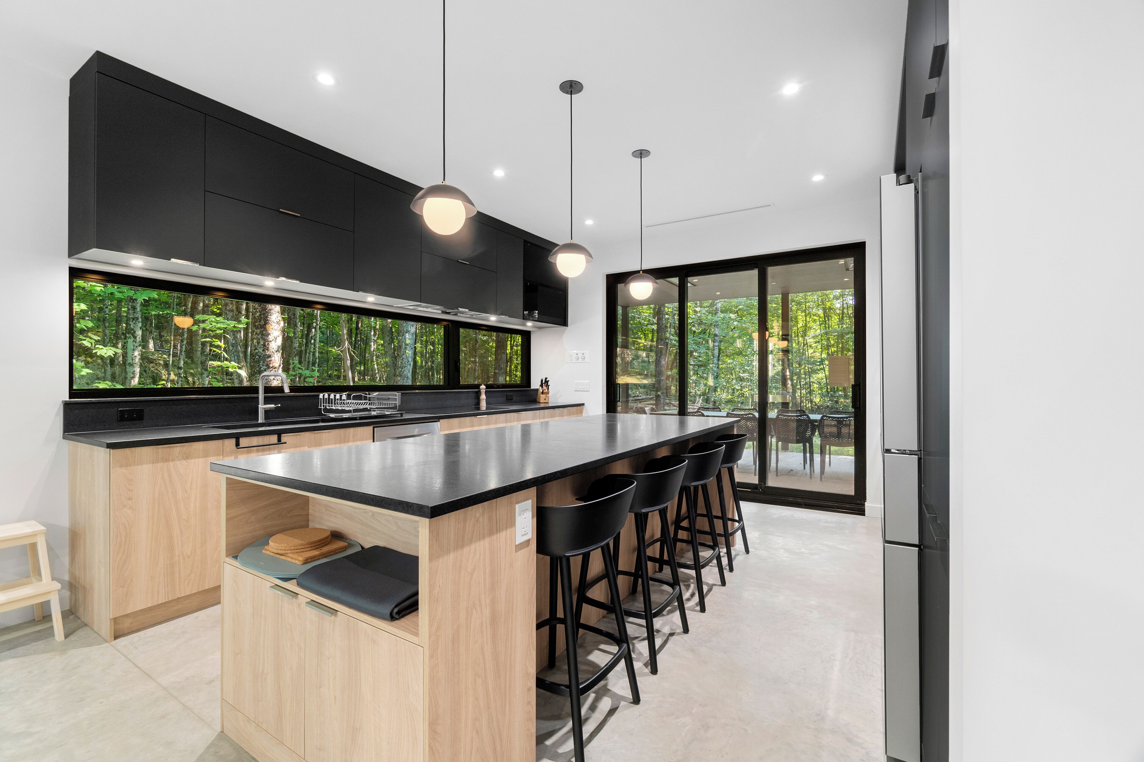 A modern kitchen with a large island, black bar stools, and a view of greenery through the glass door.