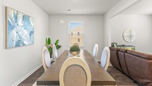 Een eethoek met een lange tafel, stoelen, een bank, een potplant en een wanddecoratie.