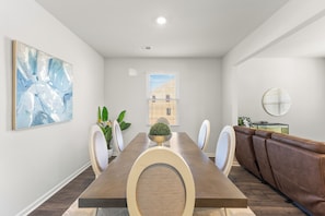 A dining area with a long table, chairs, a sofa, a potted plant, and a wall-mounted painting.