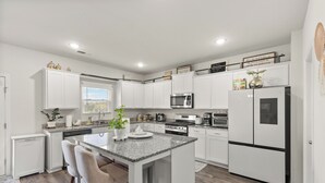 Een moderne keuken met witte kastjes, granieten aanrechtbladen, een centraal eiland met barstoelen, een koelkast, een magnetron en een fornuis.