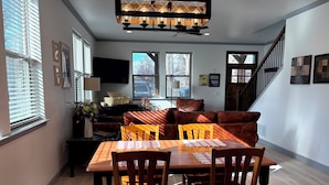 Un comedor con una mesa de madera, sillas y un sofá. Hay un televisor en la pared, una escalera y una ventana con persianas.