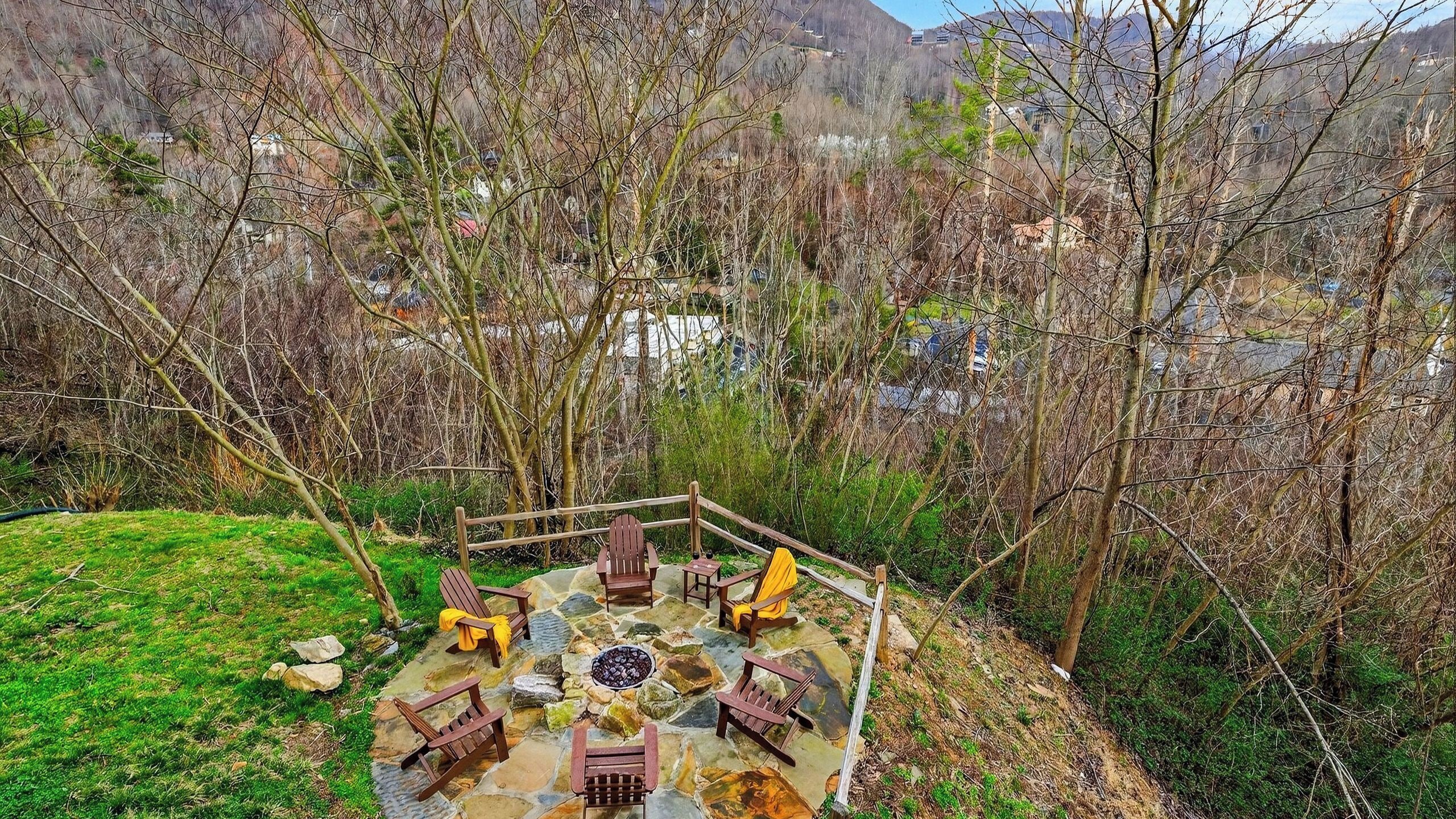 A stone deck with Adirondack chairs and a fire pit, surrounded by trees and a view of a valley.