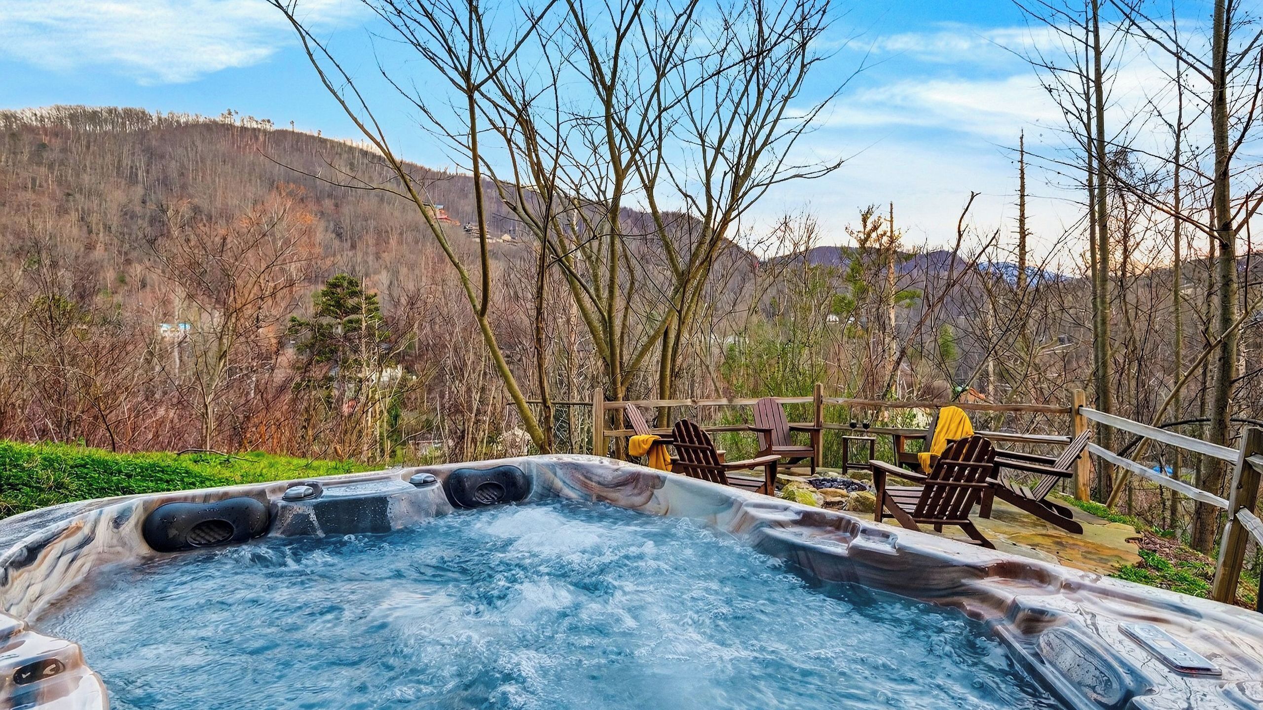 Ein Whirlpool mit Holzstühlen und einer Holzterrasse, umgeben von Bäumen und einer Bergkulisse.