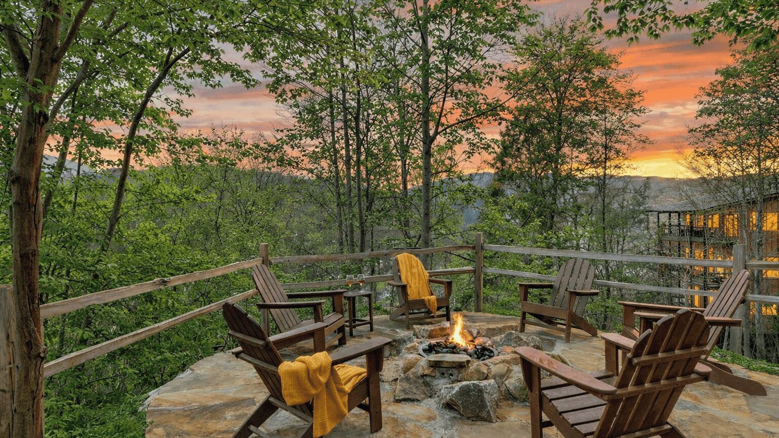 An outdoor seating area with wooden chairs and a fire pit, surrounded by trees and a sunset view.