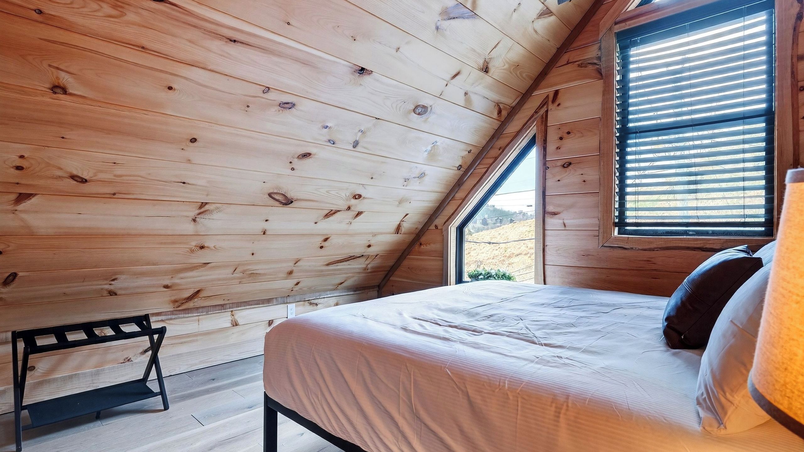 A bedroom with a sloped ceiling, wooden walls, a bed with pillows, and a window with blinds.