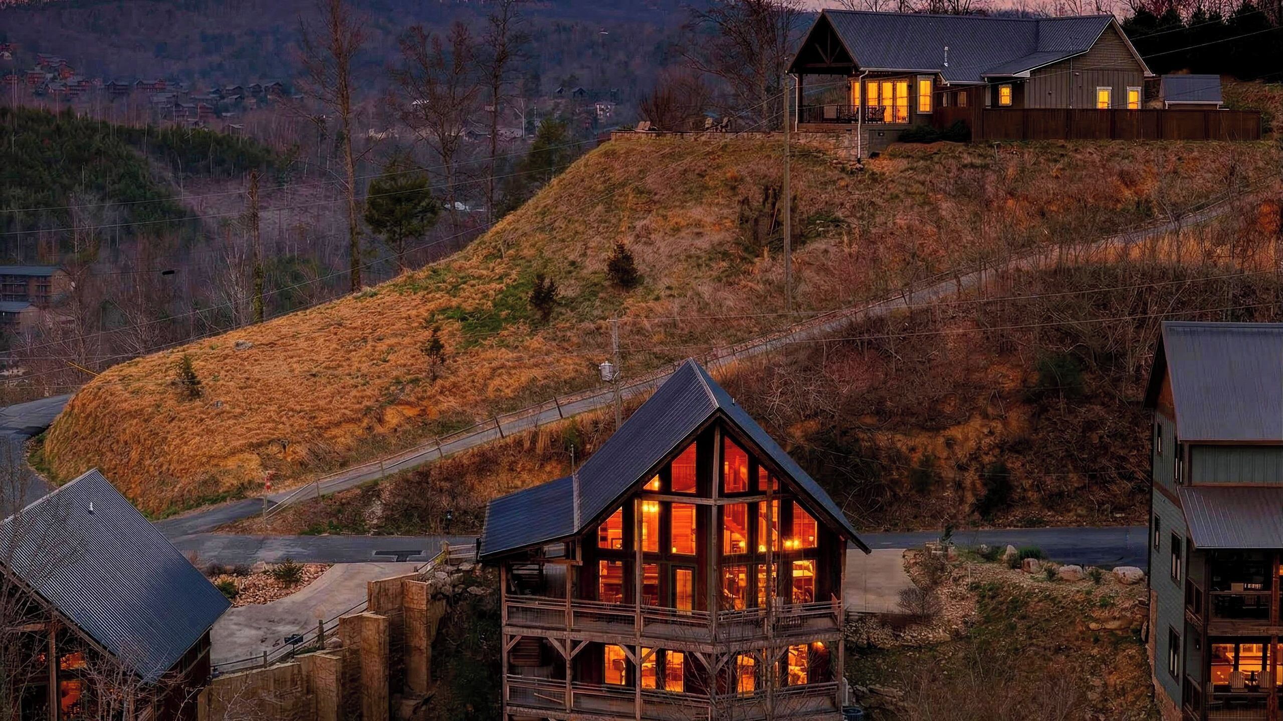 A hillside house with a prominent wooden structure and a metal roof, surrounded by a mountainous landscape.