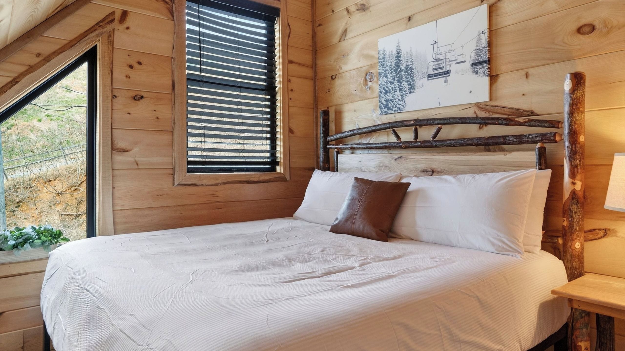 A wooden cabin bedroom with a bed, a window with blinds, and a picture on the wall.