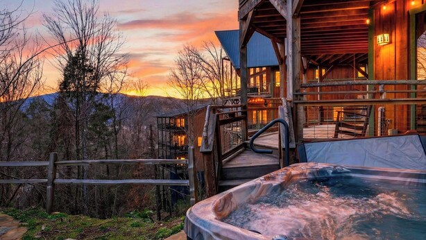 A wooden cabin with a hot tub, a wooden deck, and a view of the sunset over mountains. - Aspen Lodge by Avantstay Indoor Pool + Hot Tub + Sauna + Game Room + Mountain Views (Gatlinburg)