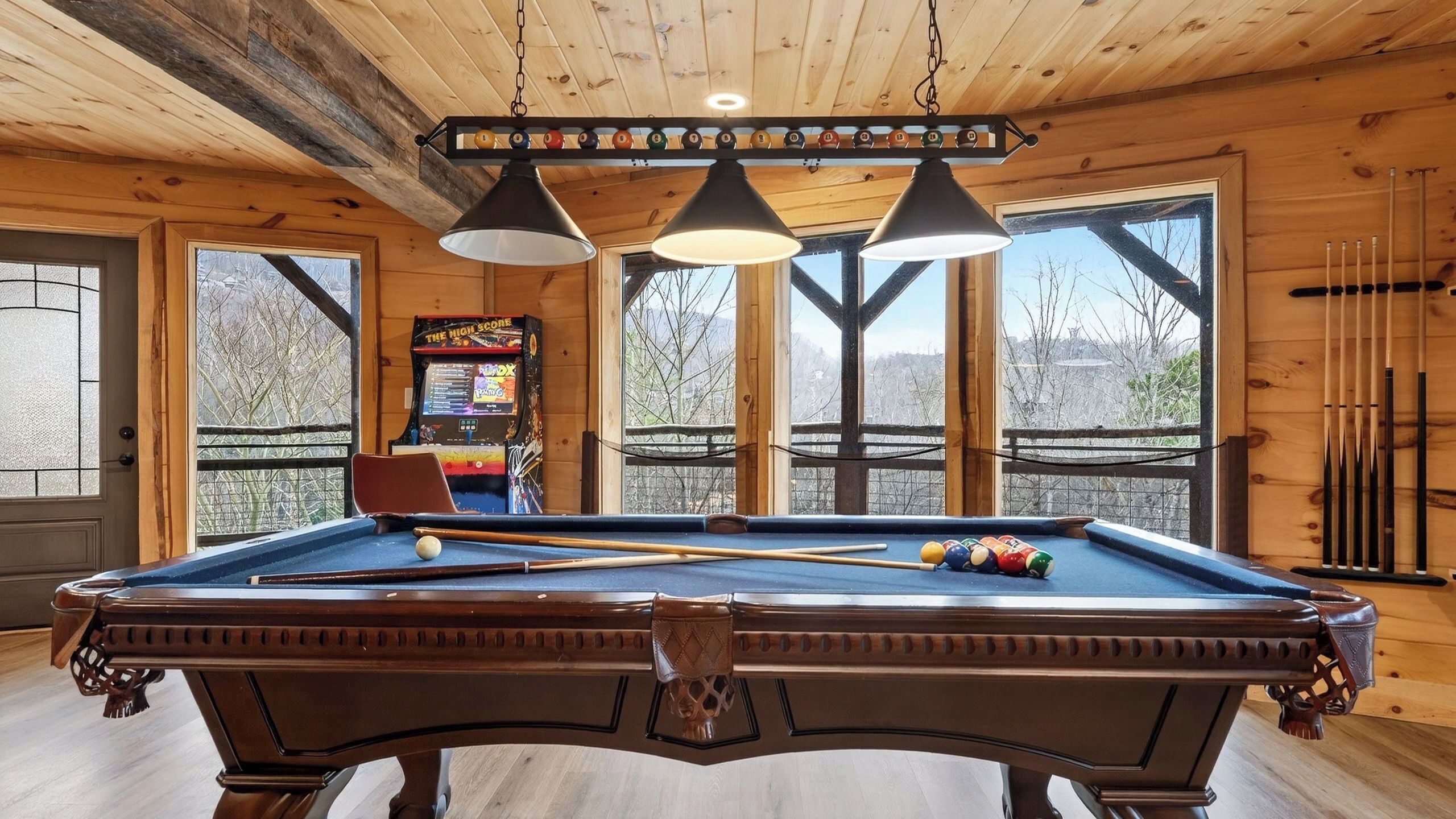 A pool table with a wooden frame, overhead lighting, and a view of a snowy landscape through large windows.