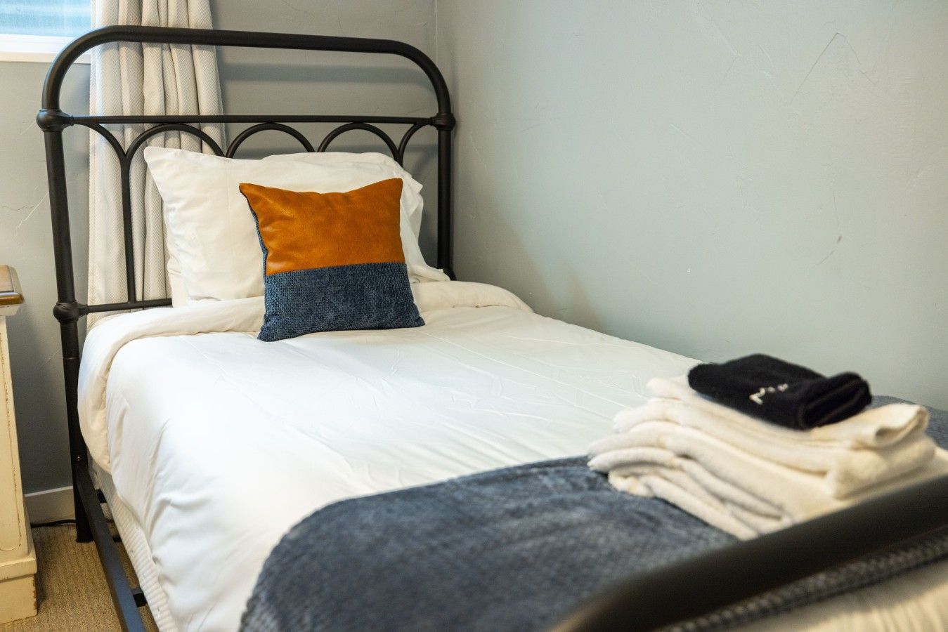 Ein Einzelbett mit Metallgestell, weißer Bettwäsche und einem blau-orangen Kissen.