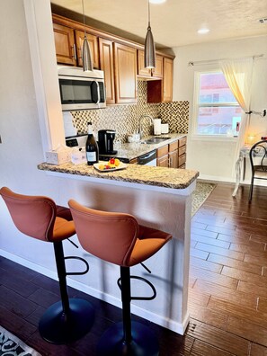 A kitchen with wooden cabinets, a microwave, a coffee maker, and a sink. There are two bar stools and a window with curtains. - Walk to OCU •Lazy Leopard Lounge •Stylish OKC Stay• 2 bedroom• KING & QUEEN (Oklahoma City)