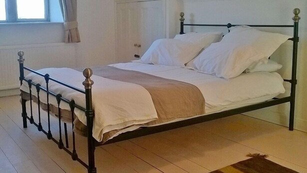 A single bed with a wooden frame and white bedding in a room with a sloped ceiling and a window with curtains. - Stable Cottage Battens Farm (Chippenham)