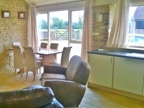 A room with a dining table, chairs, a sofa, a kitchen area, and a view of the outside. - Stable Cottage Battens Farm (Chippenham)