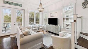 Uma sala de estar com sofá seccional, lareira e televisão. Há grandes janelas e uma placa com a inscrição 'BEACH HOUSE'.