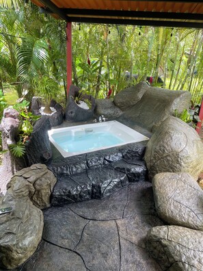 Un jacuzzi rodeado de rocas y plantas, con una estructura de madera en lo alto.