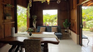 A room with a wooden table, a sofa, hanging plants, and a view of greenery outside. - Ceiba EcoContainer / La Fortuna / Jacuzzi - AC - WIFI. (Agua Azul)