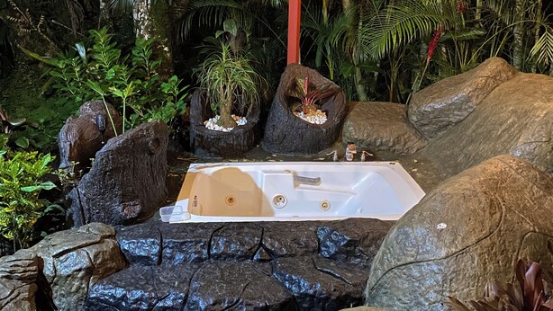 A hot tub surrounded by rocks and tropical plants, with a wooden ceiling and string lights. - Ceiba EcoContainer / La Fortuna / Jacuzzi - AC - WIFI. (Agua Azul)