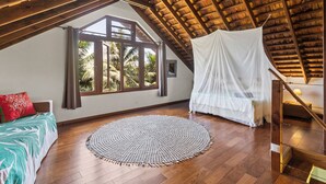 Ein Zimmer mit Holzboden, einem großen Fenster mit Blick auf Palmen, einem Bett mit rotem Kopfkissen, einem weißen Moskitonetz und einem gewebten Teppich.