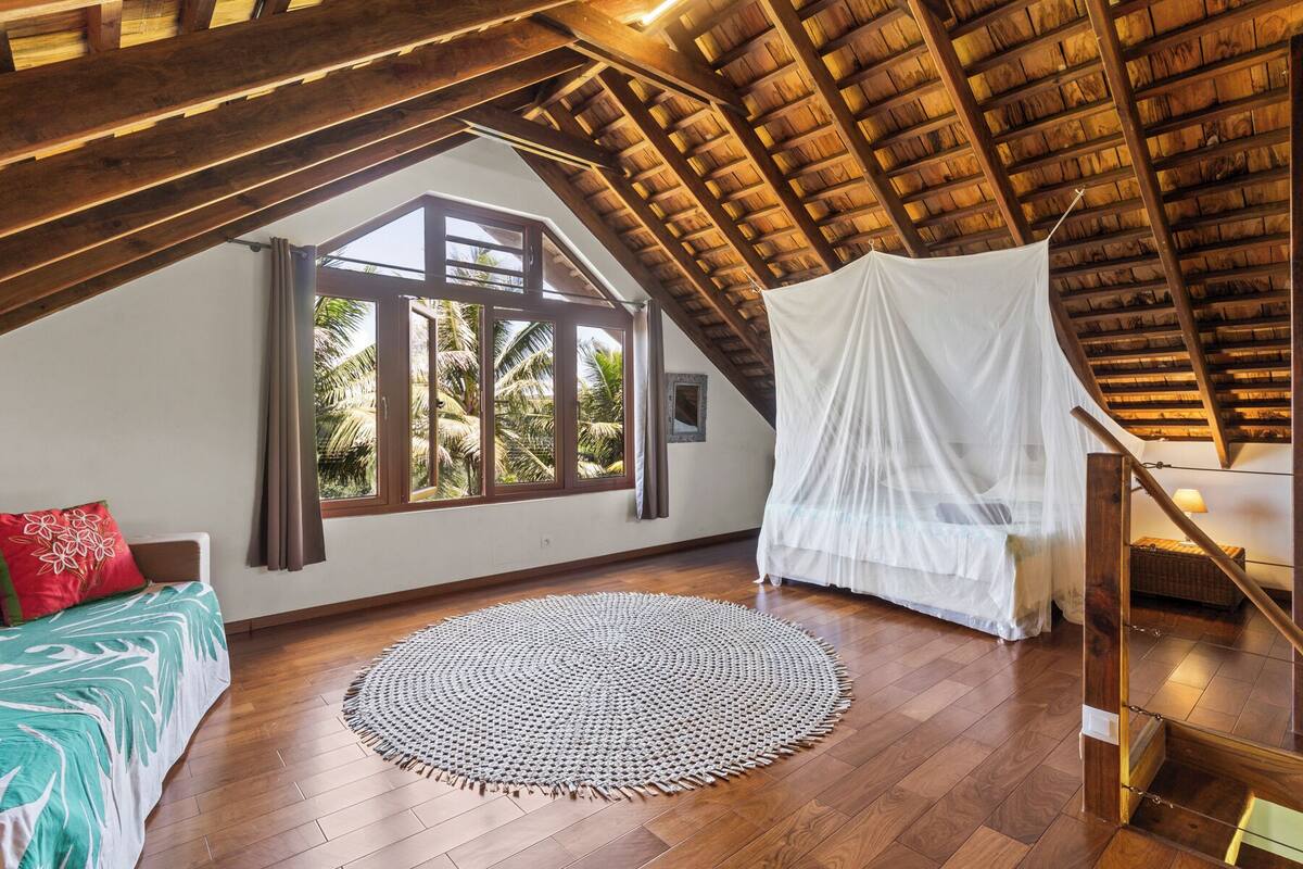 A room with a wooden ceiling, a large window with a view of palm trees, a bed with a red pillow, a white canopy, and a woven rug.
