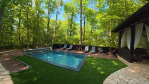 A swimming pool with adjacent lounge chairs, surrounded by a wooden deck and a stone wall, set in a forested area. - Forest home with pool, jacuzzi and sauna for relaxing and fun (Ivanec)