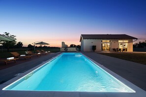 Une piscine rectangulaire à l’eau d’un bleu limpide, entourée de chaises longues et de parasols, et un bâtiment avec un toit recouvert de tuiles en arrière-plan.