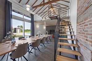 Un espace de repas moderne avec une longue table en bois pouvant accueillir six personnes, un escalier doté d’une rampe en métal et un mur en briques.
