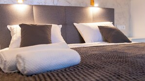 Una cama bien hecha con cabecera gris, almohadas blancas y marrones, y una toalla blanca enrollada.