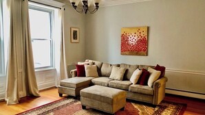 A living room with a sectional sofa, ottoman, and a chandelier. - Family friendly neighborhood spacious 3-bedrooms, 2 baths,  parking, near train. (Brookline)
