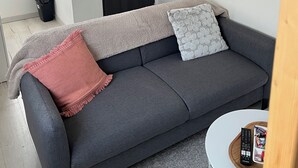 Un sofá gris con una manta y almohadillas, una mesita redonda blanca con un control remoto, una puerta negra y un adorno en la pared.