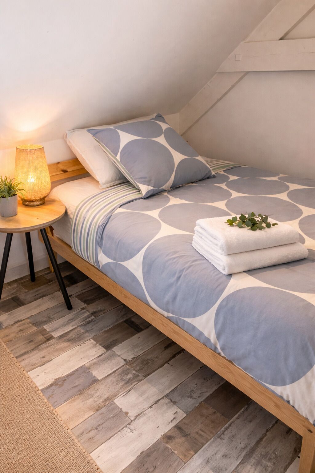 Ett sovrum med ett sluttande tak, en säng med ett geometriskt mönstrat överkast, en sänglampa, en liten växt och en hopvikt handduk på sängen.