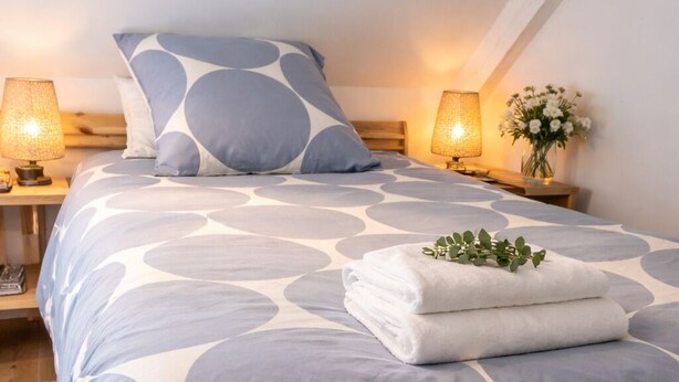Una cama bien hecha con una colcha de estampado geométrico, dos lámparas de noche, un jarrón con flores y una pila de toallas dobladas.