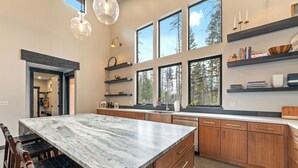 Cocina moderna con una isla grande de mármol, armarios de madera y ventanas amplias que dan a un paisaje de árboles.