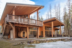 A wooden cabin with a balcony, a hot tub, and a stone wall. - New! Hot Tub, Sauna, Dog Friendly, 2 Living Spaces Near Whitefish & Glacier (Whitefish)