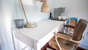 Ein weißer Schreibtisch mit einer beigen Schreibtischlampe, ein brauner Stuhl, eine Kaffeemaschine und ein Korb an der Wand.