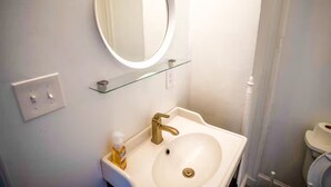 Ein Badezimmer mit einem weißen Waschbecken, einem goldfarbenen Wasserhahn und einem runden Spiegel mit Ablagefläche darunter.