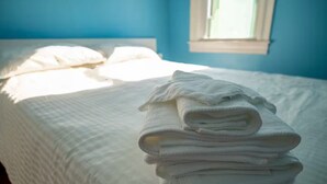 Ein ordentlich bezogenes Bett mit weißen Leinen und Kissen, ein Fenster mit einfallendem Tageslicht und eine blaue Wand.
