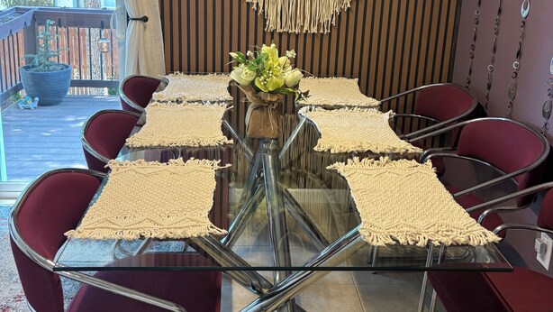 Un área de comedor con una mesa de vidrio, sillas rojas, individuales de macramé y un colgador de macramé en la pared.