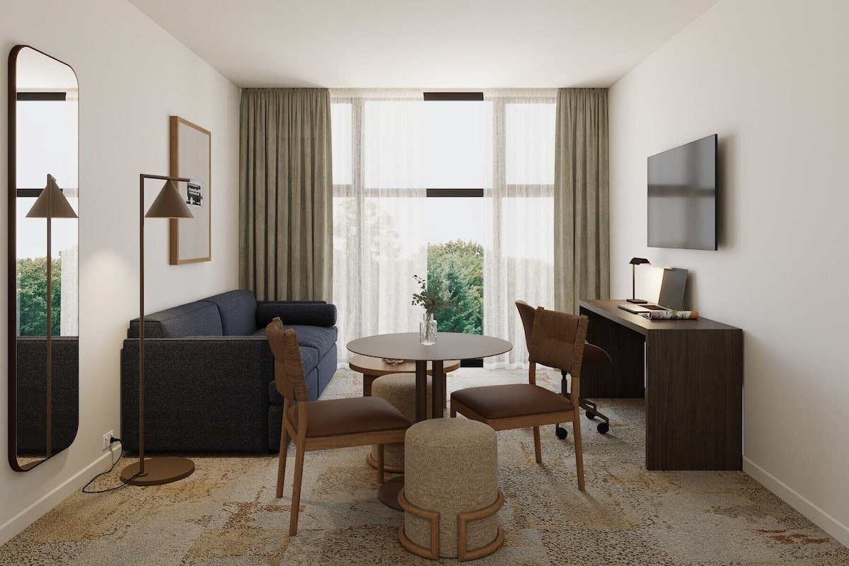 Une chambre d’hôtel moderne équipée d’un canapé, d’une table ronde, de chaises, d’un bureau, d’une télévision et d’une grande fenêtre avec des rideaux.