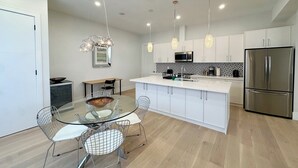 Une cuisine moderne avec des meubles blancs, un réfrigérateur en acier inoxydable, une table à manger en verre avec des chaises en fil de fer, des luminaires suspendus et un four à micro-ondes.