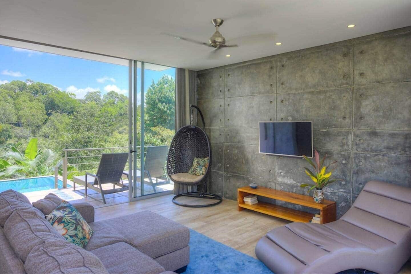 Una sala de estar moderna con un sofá, un sillón, una silla colgante, un televisor y vistas a una piscina y vegetación.