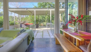 Un patio exterior moderno con sofá, mesa de comedor y sillas, rodeado de paredes de vidrio y un área cubierta.